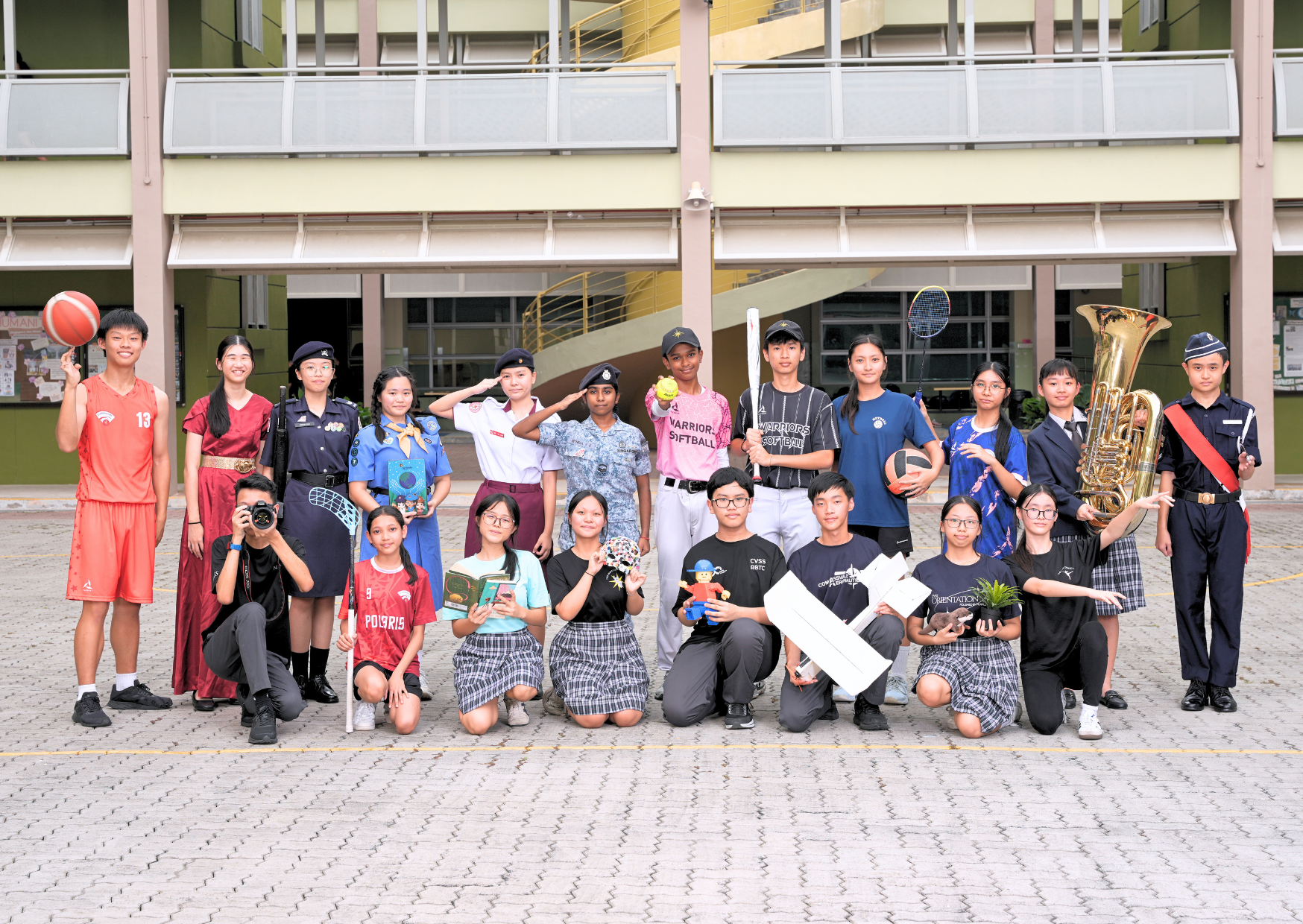 Group of school students in various uniforms and costumes holding sports equipment, books, and instruments outdoors.